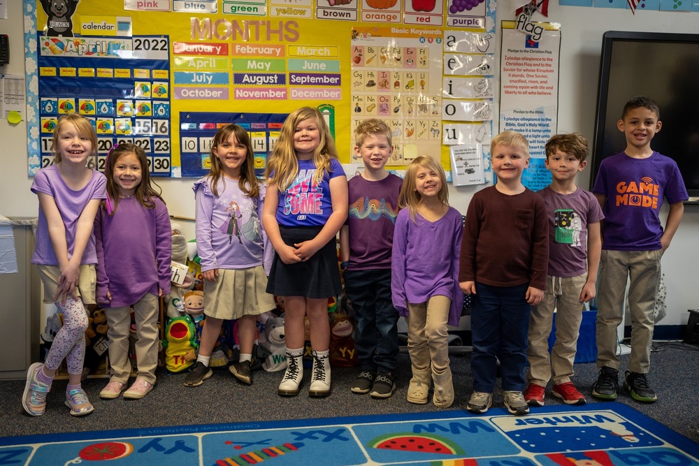 Image of nine elementary students standing in classroom wearing purple shirts. 